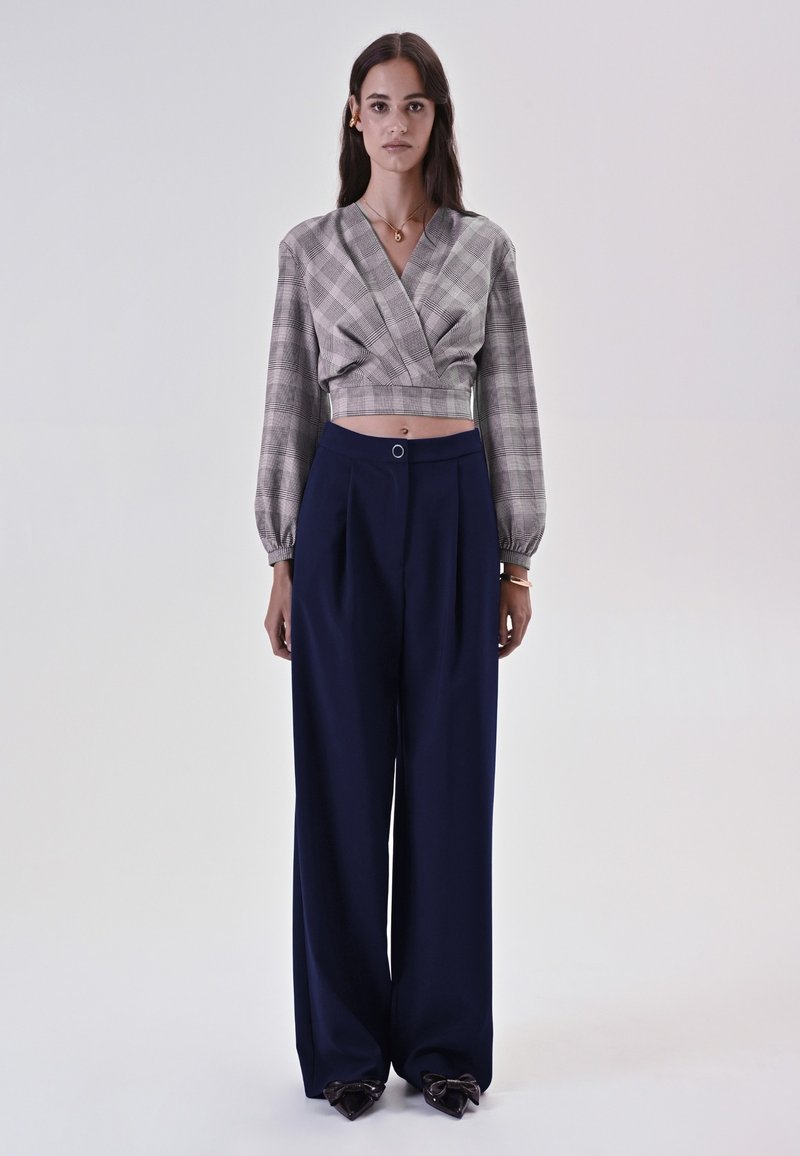 Blusa a quadri, corta, con maniche lunghe; pantaloni navy a gamba larga con pieghe; scarpe con dettagli a fiocco; gioielli semplici.