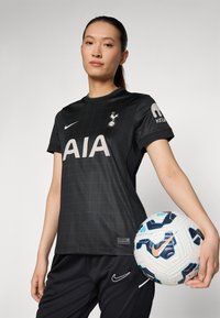 Maillot de football noir avec motif à carreaux, logo "AIA", emblème de Tottenham, associé à un short noir. Le joueur tient un ballon de football Nike multicolore.