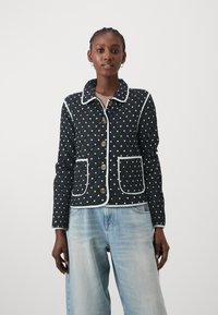 Quiltet svart jakke med hvite prikker, kontrasterende hvit piping, tre knapper og to frontlommer. Brukes over lyseblå jeans.