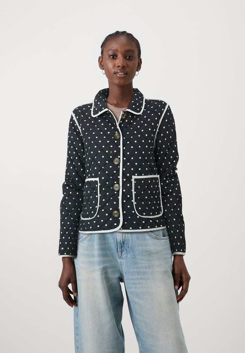 Quiltet svart jakke med hvite prikker, kontrasterende hvit piping, tre knapper og to frontlommer. Brukes over lyseblå jeans.