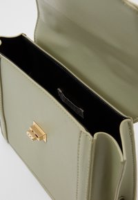 Bolso de piel sintética verde claro con un cierre metálico dorado. El interior es negro con una etiqueta. Textura suave, forma rectangular, diseño minimalista.