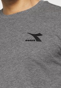 Harmaa puuvillapaita, jossa on pyöreä kaula-aukko ja musta logopainatus vasemmalla rintamuksella. Teksturoitu kangas ja rento istuvuus.