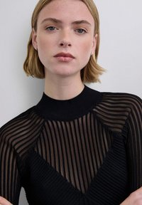 Joven con cabello rubio corto, vistiendo un top negro traslúcido a rayas, mirando directamente al frente con expresión neutral sobre un fondo liso.