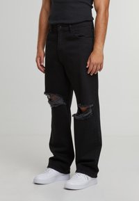 Mustad kulunud teksad, millel on suured lõiked põlvedel, valmistatud teksakangast. Sobitatud valgete tennistega, millel on sirge säärega disain.