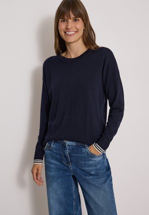 Kvinde smilende iført en mørk marineblå langærmet sweater med stribede manchetter og blå denimjeans, står foran en ensfarvet lys baggrund.