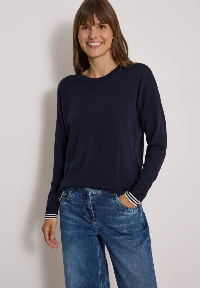Femme souriante portant un pull bleu marine à manches longues avec des poignets rayés et un jean bleu en denim, debout devant un fond clair uni.