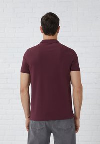 Burgundijas polo krekls ar apkakli, īsām piedurknēm un smalku faktūru, apvienots ar pelēkiem džinsu biksēm. Fons veido balta ķieģeļu siena.