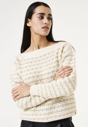 Cremfarbener Pullover mit einem strukturierten Strickmuster, verziert mit goldenen Akzenten. Off-Shoulder-Design und gerippte Bündchen, kombiniert mit dunklen Hosen.