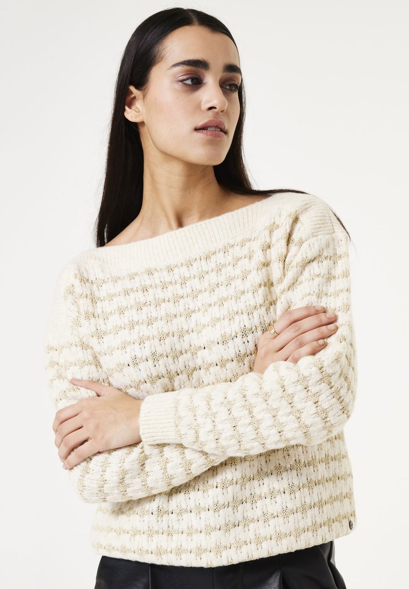 Crème trui met een textuurgebreid patroon, voorzien van gouden accenten. Off-shoulder ontwerp en geribbelde manchetten, gecombineerd met donkere broek.