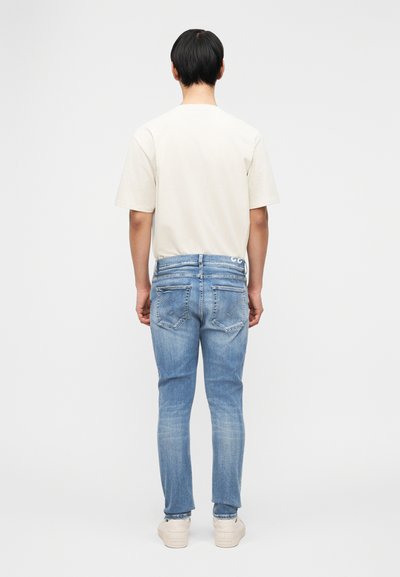 Άνδρας που στέκεται πλάτη, φοράει απλό μπεζ μπλουζάκι, ανοιχτόχρωμα τζιν και λευκά αθλητικά, με φόντο απλό λευκό.