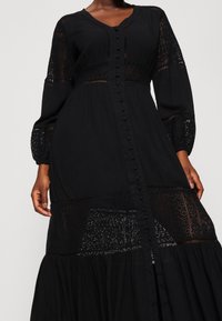 Abito lungo nero a maniche lunghe realizzato in tessuto leggero, con dettagli in pizzo, scollatura a V e chiusura frontale con bottoni, completo di una gonna fluida.