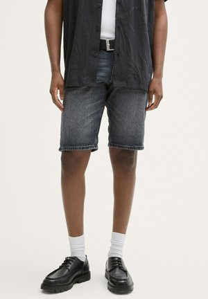 Dunkel gewaschene Denim-Shorts im klassischen Schnitt, mit geradem Bein, fransigem Saum sowie Vorder- und Hintertaschen, kombiniert mit schwarzen Schuhen und weißen Socken.
