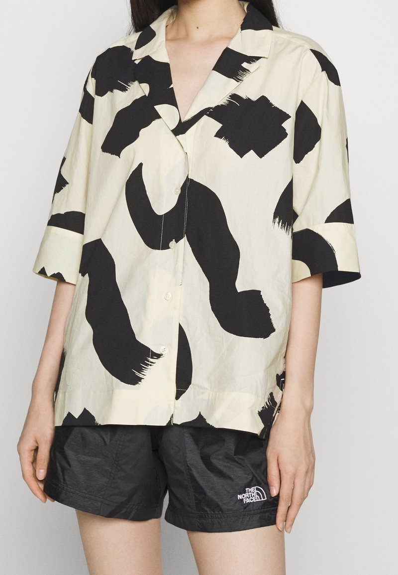 Personne portant une chemise beige avec de grandes taches abstraites noires et un short noir avec le logo The North Face sur la jambe gauche.