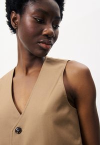 Beige ärmelloses Oberteil mit einem tiefen V-Ausschnitt, das ein einzelnes dunkles Knopfdetail und eine glatte Textur aufweist, vor einem schlichten Hintergrund.