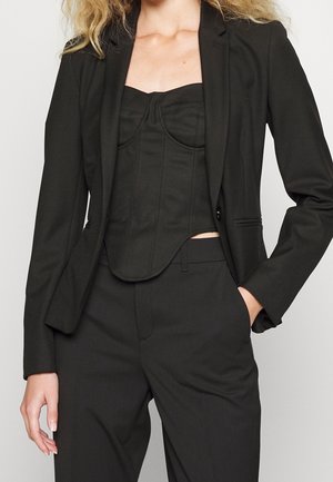 Vrouw draagt een zwarte, op maat gemaakte blazer, zwart korsetstijl topje en bijpassende zwarte hoge taille broek, poseert met één hand in de zak.