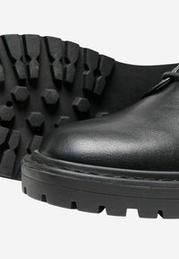 Botín de cuero negro con punta redondeada, que presenta una suela de goma negra texturizada con un patrón hexagonal y detalles de costura visibles.