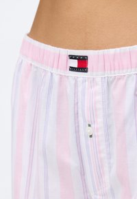 Primer plano de la cintura de pantalones de pijama a rayas en rosa claro y blanco, con parche con el logo de Tommy Hilfiger y un botón blanco en la parte delantera.