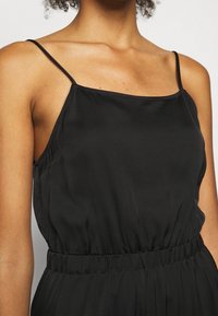 Jumpsuit sans manches noir en tissu léger ; il présente un décolleté carré, une taille élastique et une texture fluide.