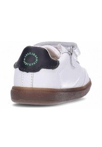 Zapatillas blancas para niños con suela marrón y parche negro en el talón, con texto circular verde y correa de velcro.