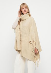 Poncho em malha bege com um colarinho alto, apresentando tecido texturizado e acabamentos em franjas ao longo das bordas, combinado com calças de cor clara.