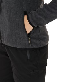 Veste grise zippée avec texture horizontale, poche latérale et bord noir, associée à un pantalon noir avec un petit logo et des détails de poche.