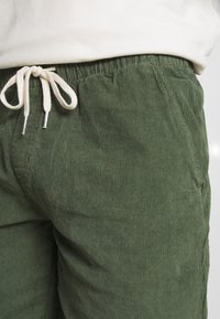 Pantalon en velours côtelé vert avec une surface texturée, doté d'une taille élastique avec un cordon de serrage beige et des accents métalliques argentés.