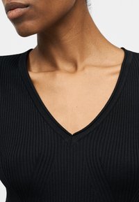 Κοντινό πλάνο ενός ατόμου που φοράει μαύρη ριμπ μπλούζα με λαιμόκοψη σε σχήμα V, εμφανίζοντας τον λαιμό, την κλείδα και μέρος του κάτω μέρους του προσώπου με φόντο ένα απλό υπόβαθρο.