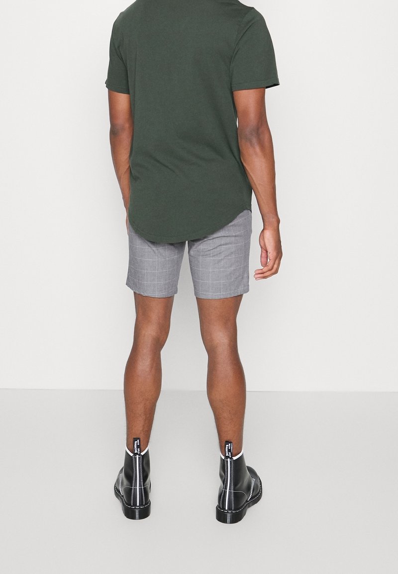 T-shirt vert foncé à manches courtes associé à un short gris à carreaux. Le short a une coupe décontractée et est fabriqué dans un tissu léger. Des bottines noires complètent le look.