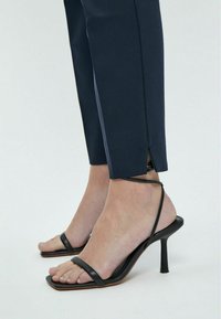 Sandali neri con tacco alto e cinturino sottile alla caviglia, caratterizzati da una punta quadrata, abbinati a pantaloni sartoriali blu scuro con spacco laterale.