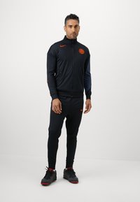 Fekete sportos melegítő, cipzáras dzsekivel és hozzáillő nadrággal. Narancssárga Nike logóval, slim fit szabással és cipzáras zsebekkel rendelkezik. Texturált anyagból készült.
