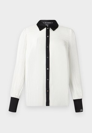 Blouse blanche plissée avec col et poignets noirs, fermeture à boutons sur le devant, et manches longues. Texture lisse avec un léger éclat.