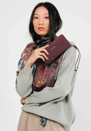 Cartera burdeos hecha de un material texturizado, con cierre de cremallera y logotipo en relieve, sostenida contra una bufanda estampada y un suéter gris.