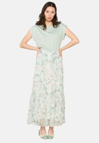 Svetlo zelena kratka majica v kombinaciji s pastelno cvetlično maksi krilo, ki ima mehke naborke, in meri z metinamesnatimi blokovnimi petami.