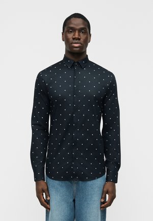 Čierna košeľa s dlhým rukávom na zapínanie na gombíky s malým bielym geometrickým vzorom, ktorú má na sebe stojaci muž v modrých džínsoch na bielom pozadí.