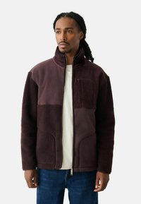 Maroon fleece jacket with contrasting textured panels and a zip front. Features a high collar and pocket on the chest. Smooth and soft texture.