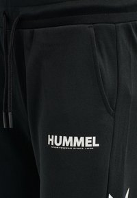 Zwarte sweatpants met een gestructureerde afwerking, voorzien van een wit "HUMMEL" logo en een tailleband met trekkoord. Inclusief zijzakken voor functionaliteit.