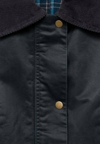 N�ärbild av svart jacka med kordflätsad krage, metallknappar och en blå rutiga skjorta synlig vid halsringningen.