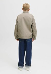 Person med kort blont hår som bär en beige jacka, mörkblå vida jeans uppvikta vid vristerna och vita sneakers, står med ryggen vänd framåt.