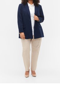 Marinblazer med två framfickor, bärs över en vit skjorta, ihop med beige byxor och nude högklackade skor. Slät tygstruktur.