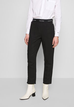 Persona con camisa blanca de manga larga metida dentro de pantalones negros Calvin Klein con cintura con logo y botas blancas de tacón hasta el tobillo, de pie sobre un suelo gris.