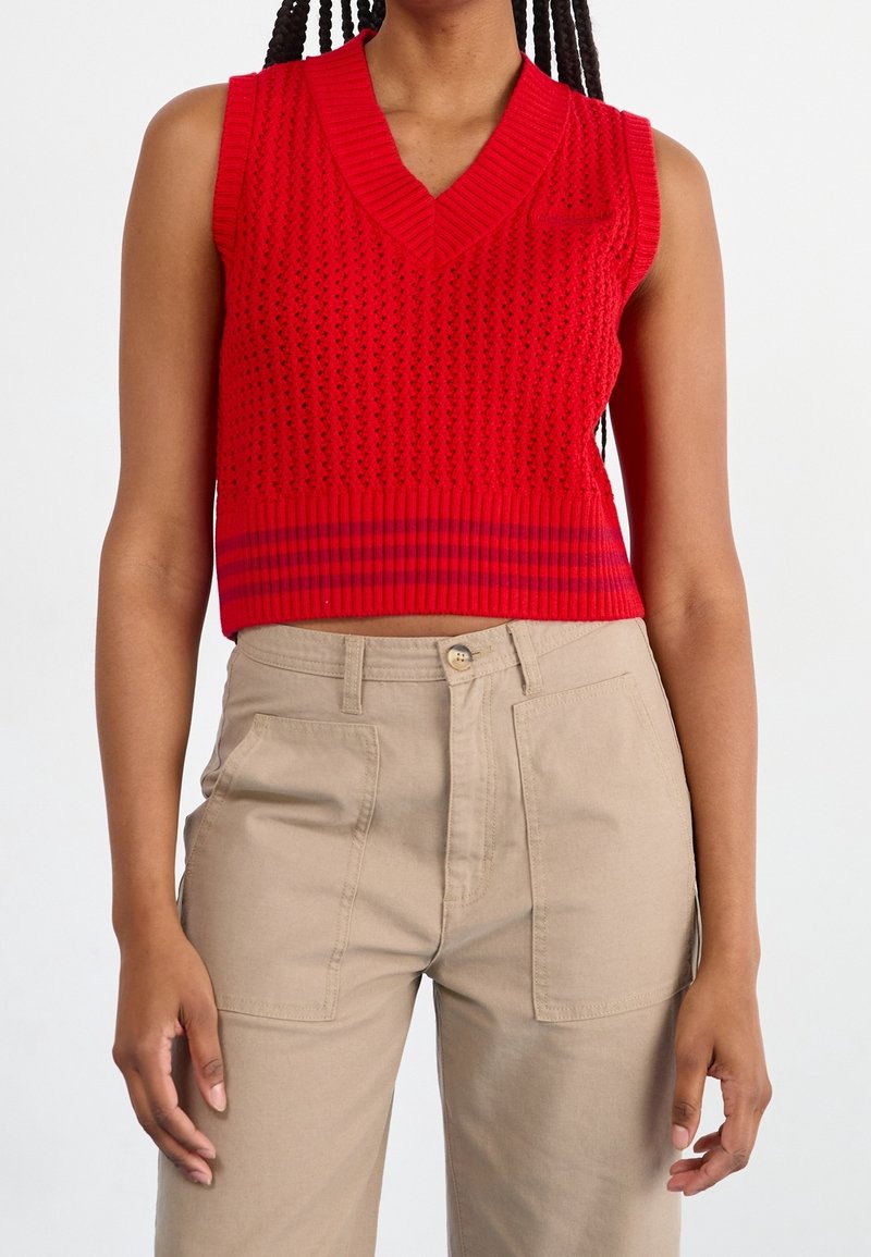 Rode gebreide mouwloze top met een v-hals, voorzien van een gestructureerd patroon en een gestreepte ribbenzoom, gecombineerd met beige hooggetailleerde broeken.