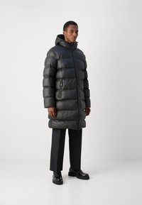Zwarte pufferjas met een capuchon, voorzien van een glanzende textuur, zijzakken met rits en een ontwerp tot op de knie, gedragen met zwarte broeken en schoenen.