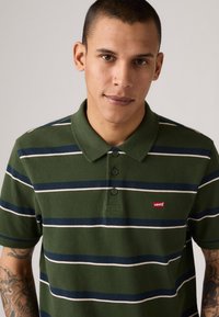 Zelené polo tričko s pruhmi, ktoré má námornícke a biele horizontálne pruhy, dvojitý gombíkový zapínanie a malé červené logo Levi's na hrudníku.