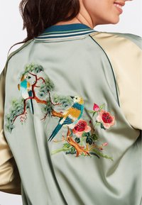 Giacca bomber in raso con base verde pastello, caratterizzata da uccelli e fiori colorati ricamati sul retro e maniche beige a contrasto.