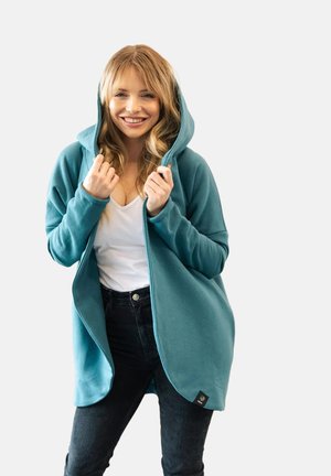 Kobieta uśmiechająca się i podciągająca kaptur tealowej kurtki, ubrana w biały top i czarne dżinsy, stojąca na jasnym, jednolitym tle.