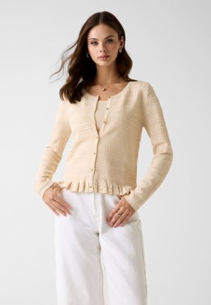 Femme portant un cardigan beige texturé avec ourlet volanté sur un haut assorti, associé à un pantalon blanc taille haute, posant devant un fond uni.