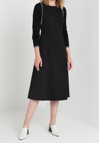 Svart långärmad klänning med en utsvängd kjol och dekorativ silverstygn längs axlarna, kombinerad med vita klackskor med blockheel.
