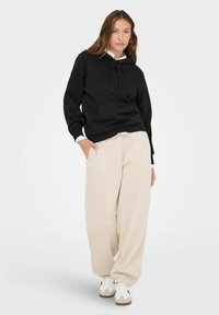 Czarna bluza z kapturem z kieszenią z przodu, w zestawieniu z jasnobeżowymi spodniami dresowymi. Sneakersy mają czarno-białe akcenty. Prosty, casualowy styl.