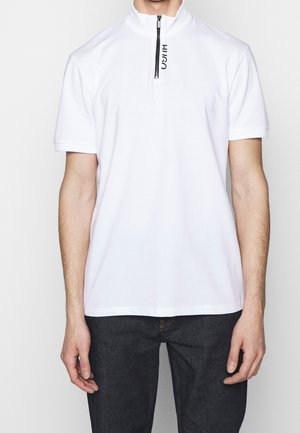 Mann trägt ein weißes Polohemd mit kurzen Ärmeln, schwarzem Reißverschluss und "HUGO"-Schriftzug in der Nähe des Kragens, kombiniert mit dunklen Jeans.