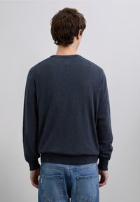 Ciemnoszary sweter z długimi rękawami i prążkowanymi mankietami, z okrągłym dekoltem, wykonany z miękkiego materiału, noszony na niebieskich dżinsach.